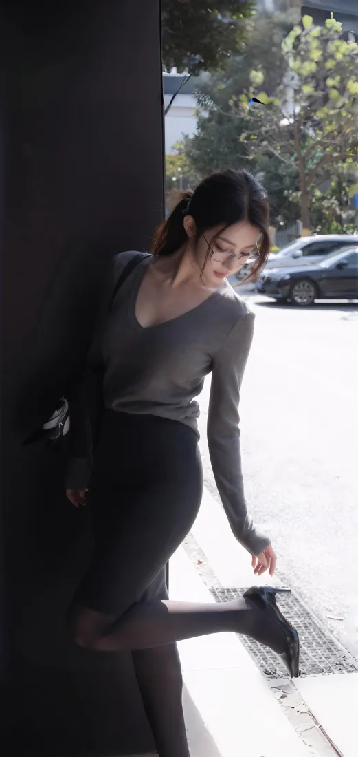 户外街边，灰衣马尾女子靠立柱抬腿整理鞋履，背景有树木与车辆