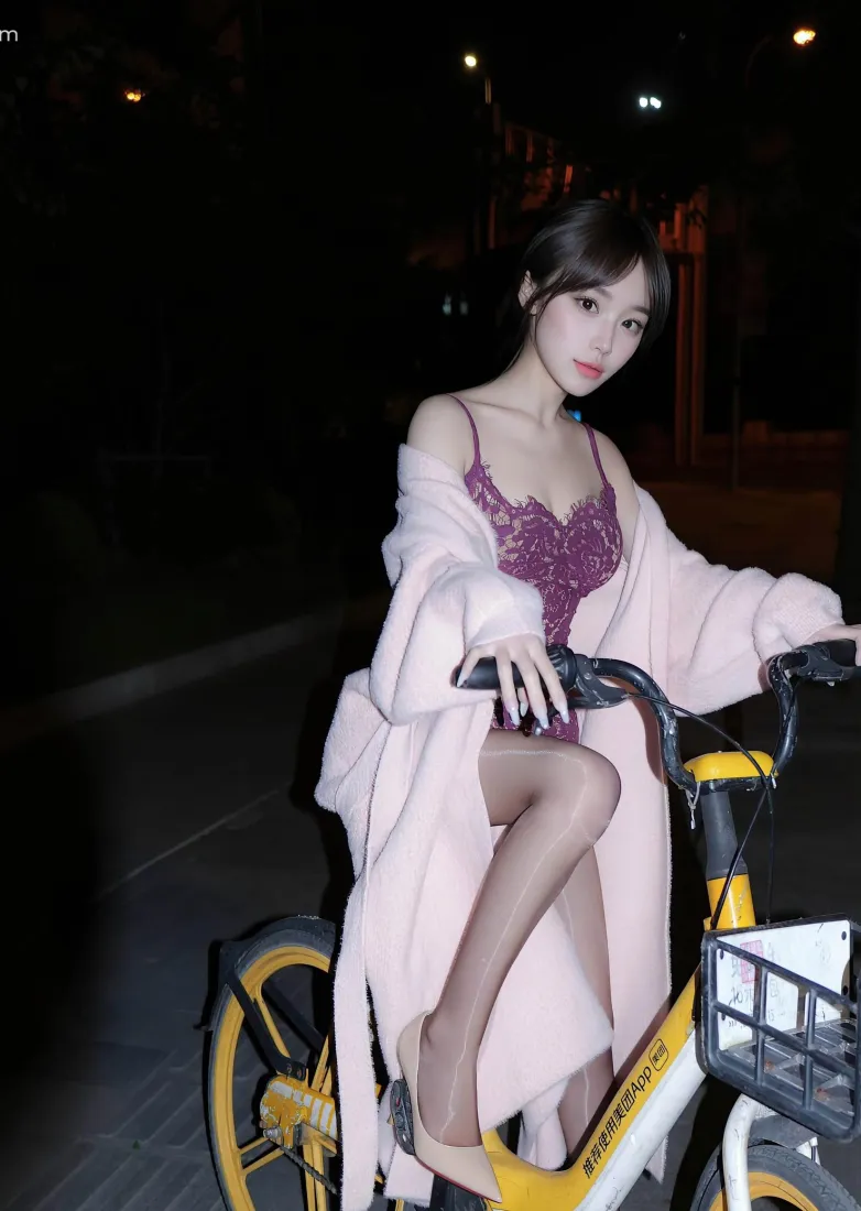 夜晚街头，穿粉色外套紫色吊带的女子倚靠在黄色共享单车旁。