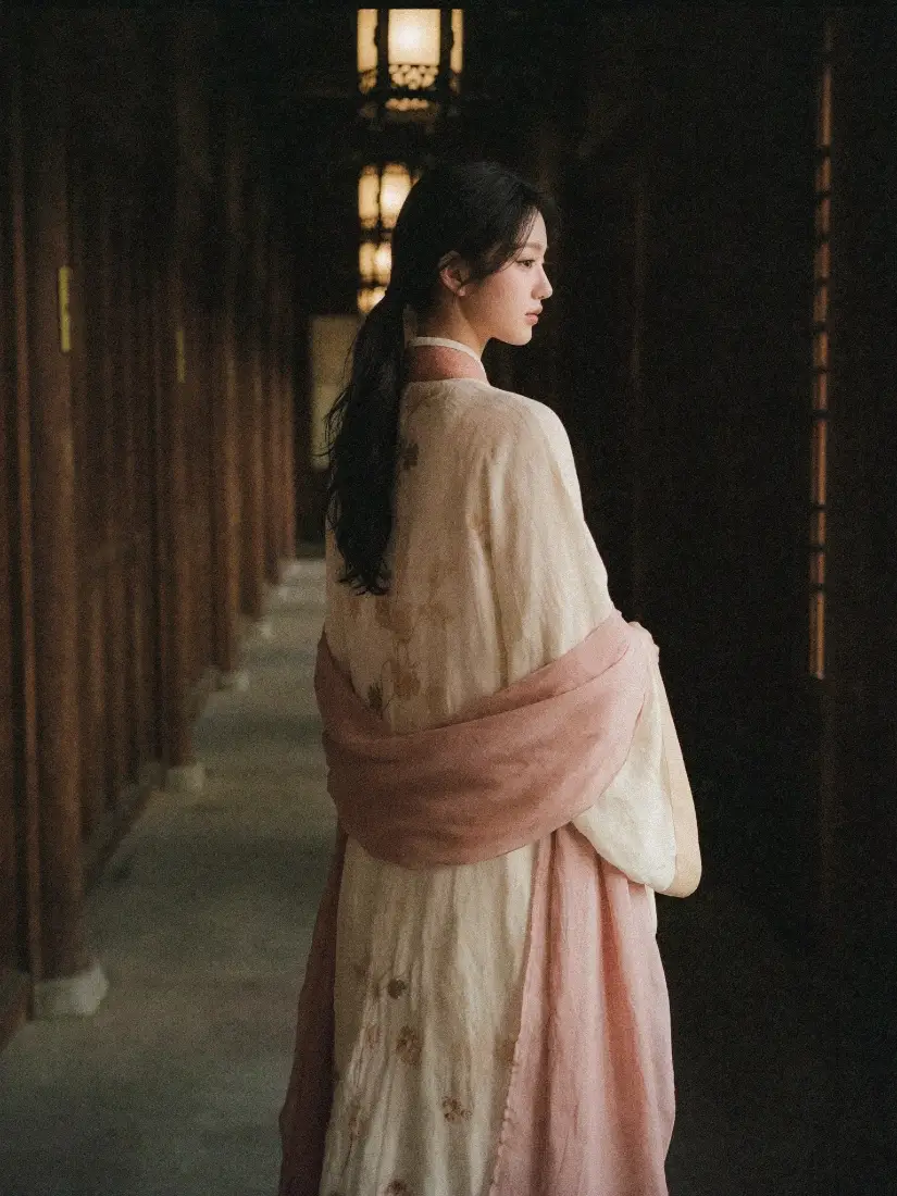 身着粉白汉服的女子立于古建木质长廊中，侧身回望，氛围温婉暗调。