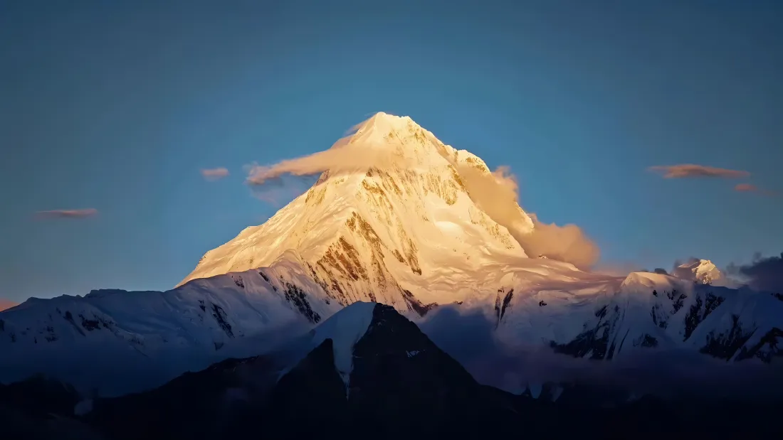 澄澈蓝天之下，日照金山的雪山主峰被暖金色阳光照亮，下方是覆雪的连绵群山。