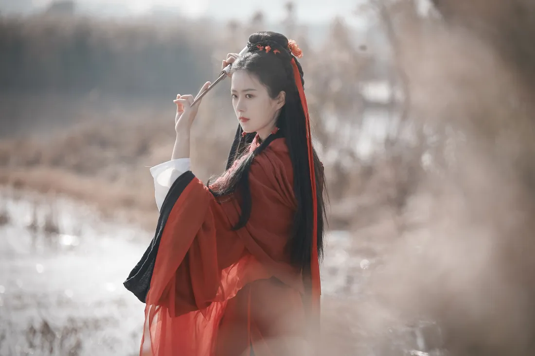 红衣汉服女子立于水边荒草间，手持长笛，神情怅然，背景朦胧虚化。