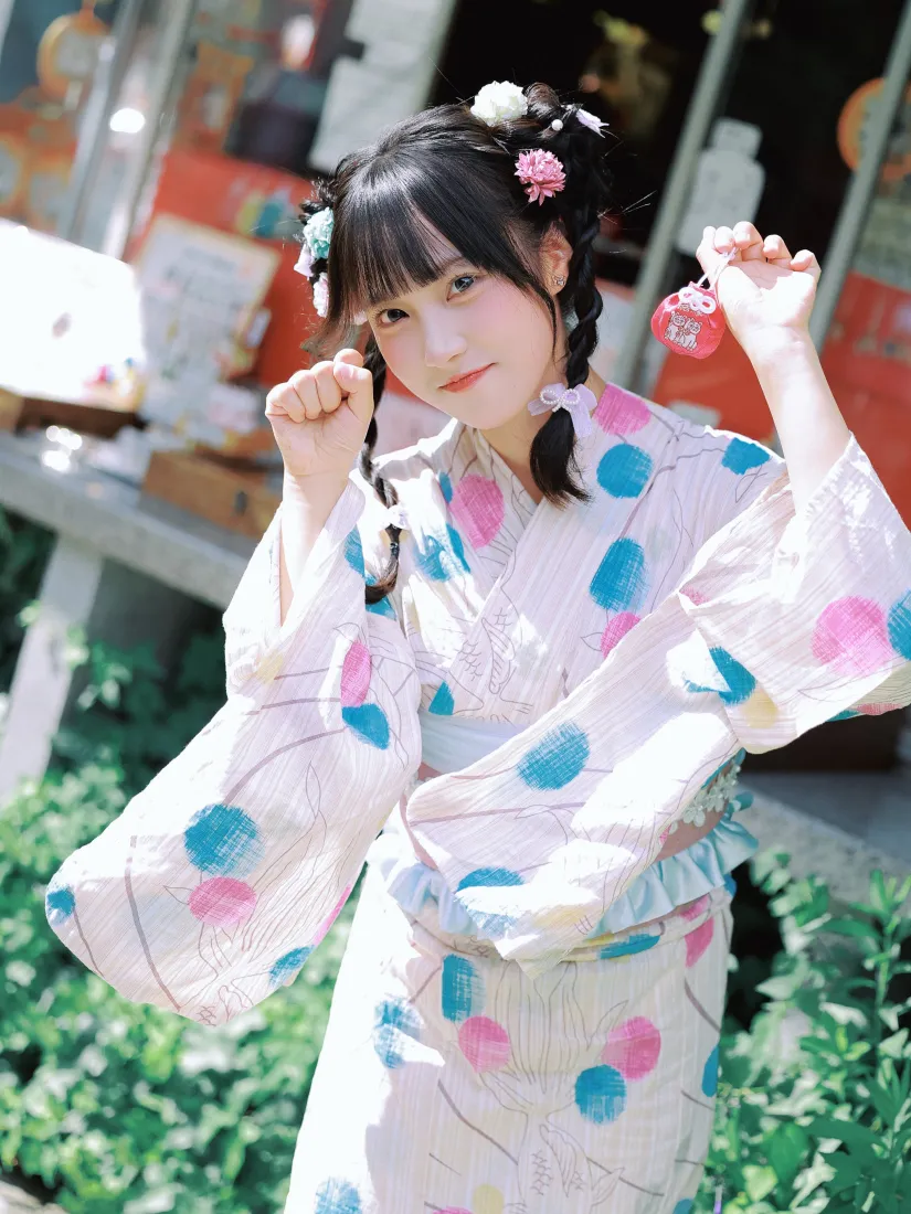 户外绿植旁，穿粉蓝波点和服的双马尾少女俏皮抬手，背景虚化。