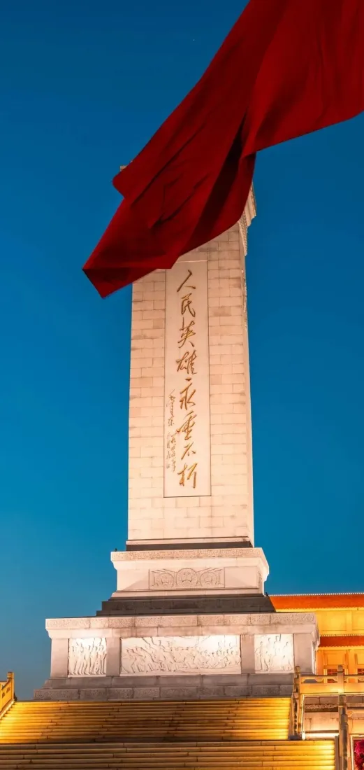 仰视视角下的人民英雄纪念碑，上方有飘动的红旗，背景是澄澈蓝天。