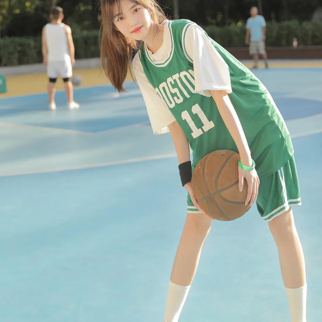 穿绿色11号篮球服的少女手持篮球站在户外篮球场，背景有模糊行人和绿植。