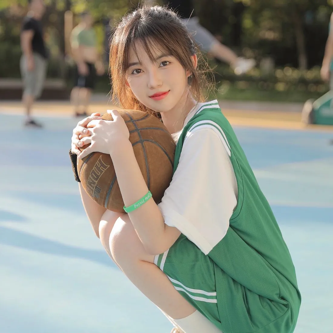 穿绿色篮球服的少女蹲在户外篮球场，双手抱篮球，背景虚化有树木和行人。