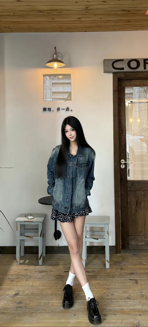 长发女生站在咖啡馆内，穿牛仔外套配碎花短裙，双手插兜双腿交叉站立。