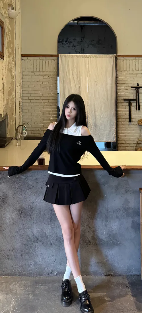 黑长直少女身着露肩黑上衣与百褶裙，倚靠在室内吧台前，姿态舒展。