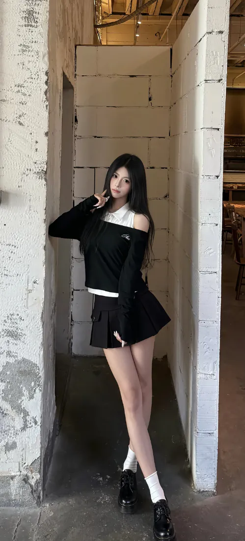 长发少女身着黑白露肩上衣与百褶短裙，在工业风室内立柱旁摆拍。
