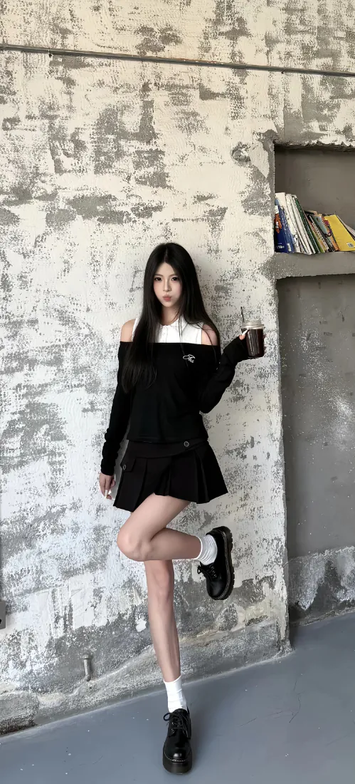 黑长直少女穿黑白露肩套装，单腿抬起手持咖啡杯，靠斑驳水泥墙站立。