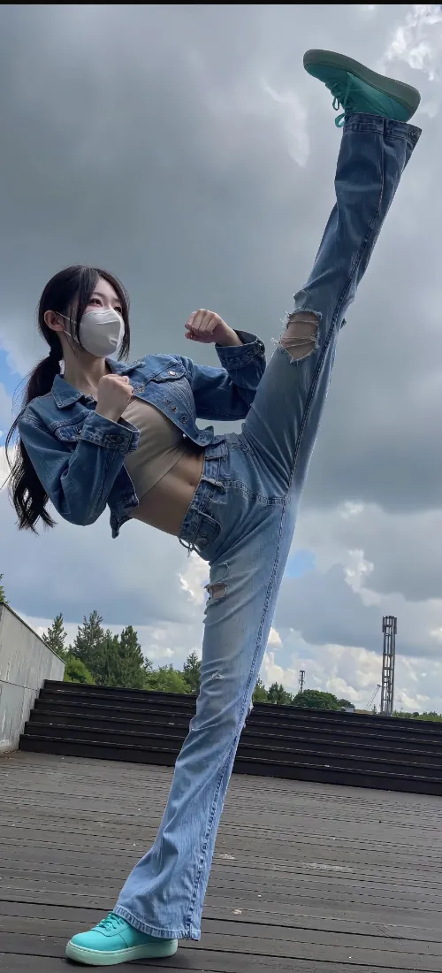 戴口罩的长发女生身着牛仔套装，在户外阴天环境下做出高踢腿动作。