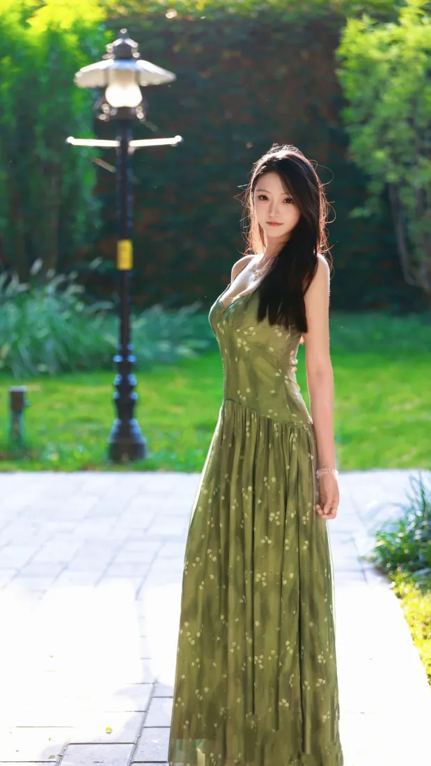 长发女子身着绿色碎花吊带长裙，站在公园步道上，背景是草坪、路灯与葱郁绿树。