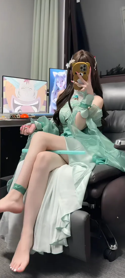 穿浅绿色露肩纱裙的长发少女坐在电竞椅上，用手机挡脸自拍，身后摆放双显示器。
