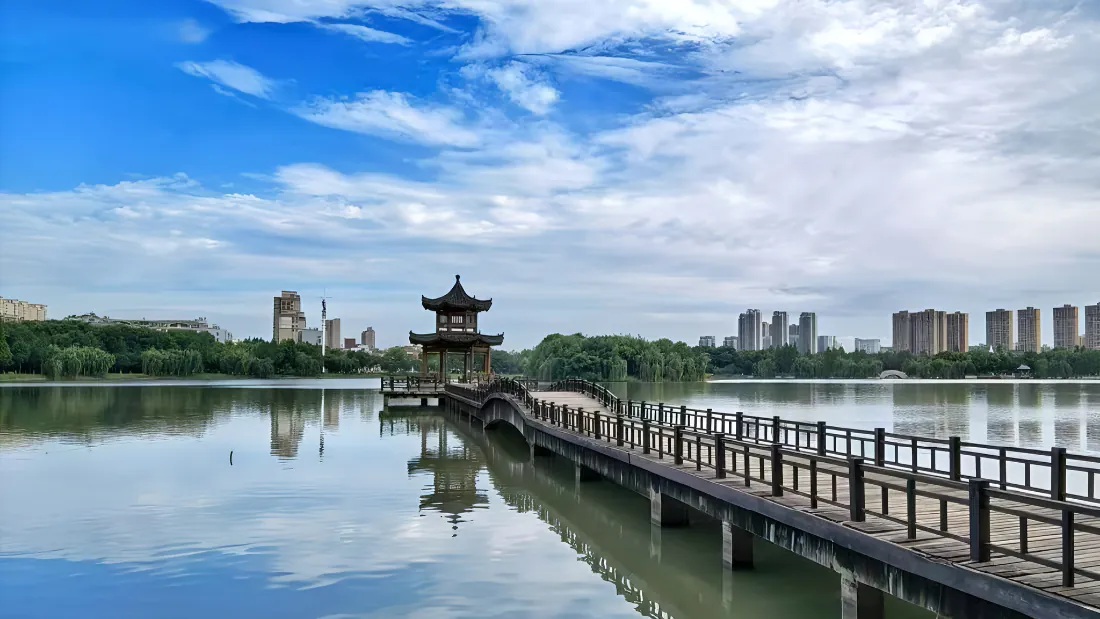 2k湖景壁纸|湖心亭木栈道湖岸背景图片 - 清新城市风光「哲风壁纸」 平静湖面延伸着木栈道,尽头立着中式古亭,远处绿树与城市高楼相映,蓝天白云倒映水中。