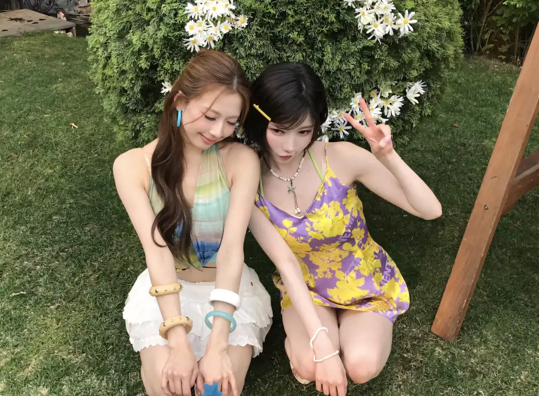 两名年轻女生蹲在草坪上，身后是开满白色小花的绿篱，夏日清新户外写真。
