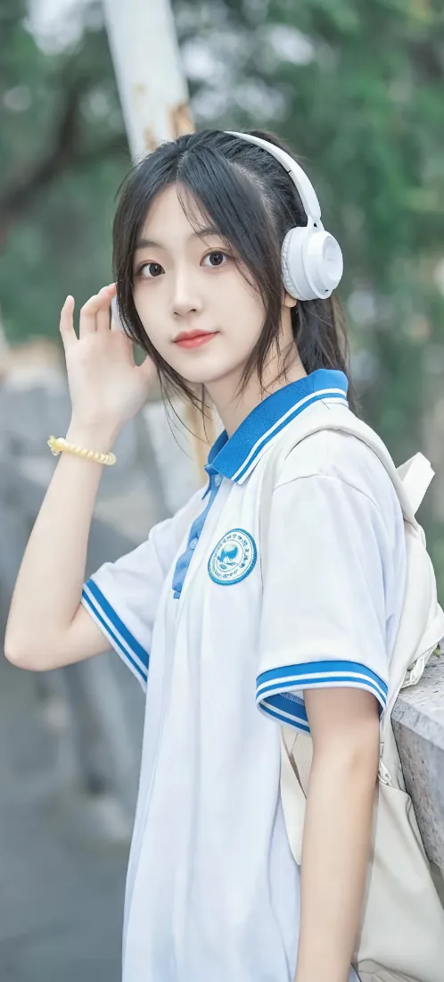 户外虚化绿植背景前，穿蓝白校服的长发少女戴白色耳机抬手扶发。