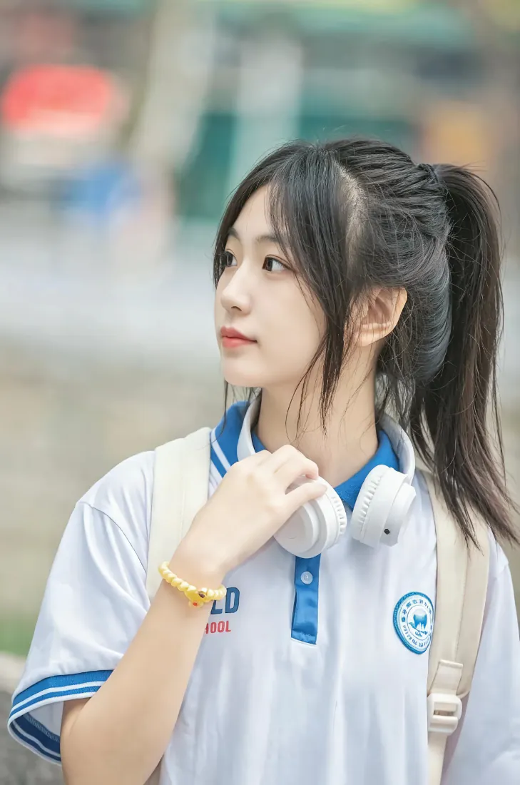 户外虚化背景前，穿蓝白校服扎高马尾的少女手扶颈间白色头戴耳机。