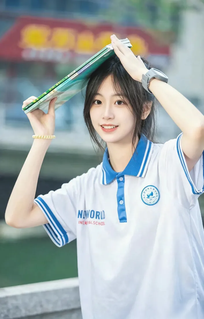 穿白蓝校服的少女双手举书本置于头顶，面带甜笑，背景虚化的校园户外场景。