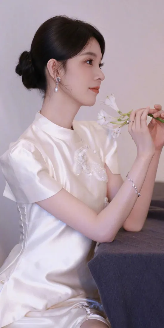 陈瑶身着米白缎面旗袍，低盘发佩戴珍珠耳饰，手持白花侧身倚靠桌沿，气质温婉清雅。