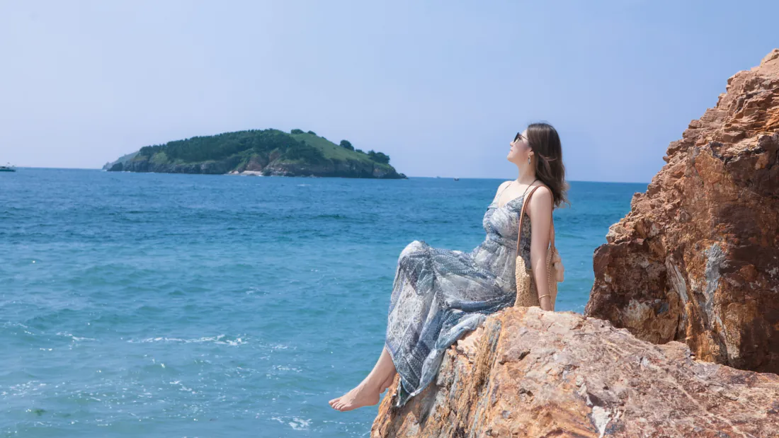 穿吊带长裙的女子赤脚坐海边礁石上，面朝碧海蓝天与远处海岛。