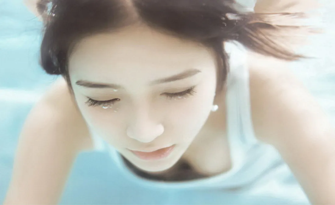 水下浅蓝环境中，穿白背心的长发少女闭眼，双手扶头，氛围柔和治愈。