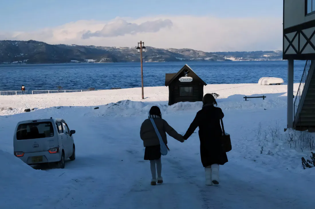 冬日雪地上两名女性牵手走向蓝色海边，远处是覆雪的连绵山脉。