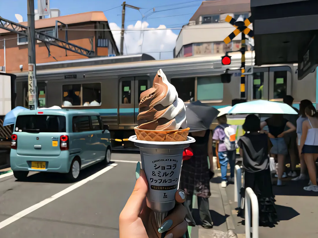 晴天日本铁路道口，前景手持双色冰淇淋，背景电车、微型面包车与等候行人。