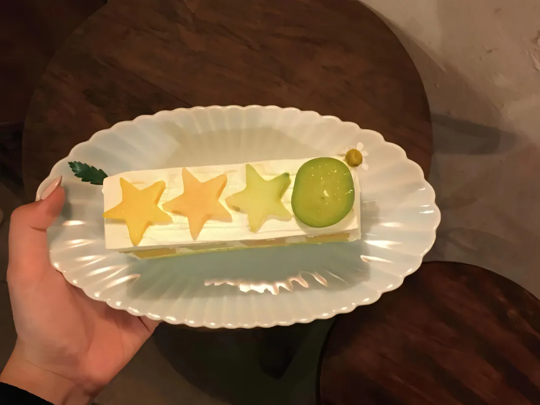 一只手托着花边白瓷盘，盘中长条奶油蛋糕饰有三色星星水果片与青提。