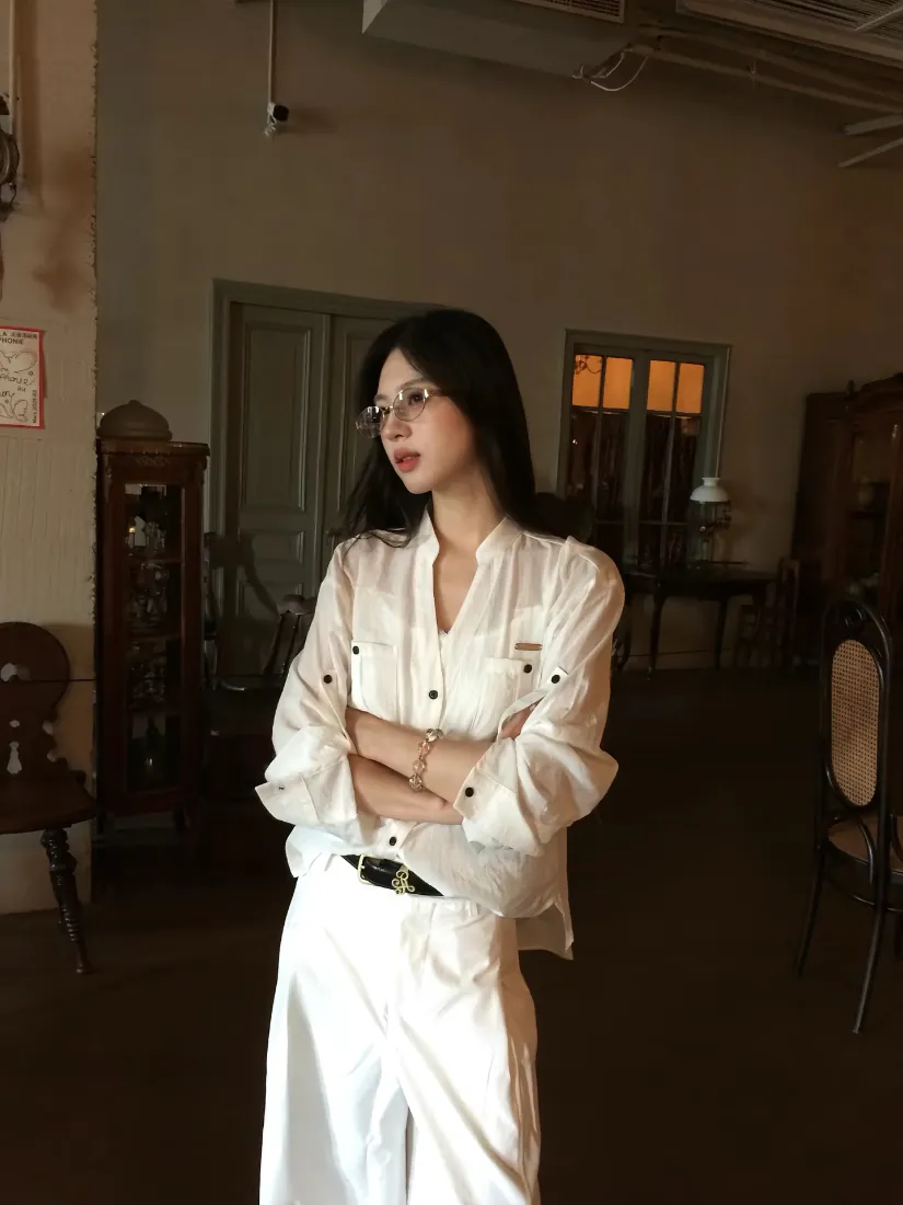戴细框眼镜的长发女子身着白色套装抱臂站在复古旧室内空间中。