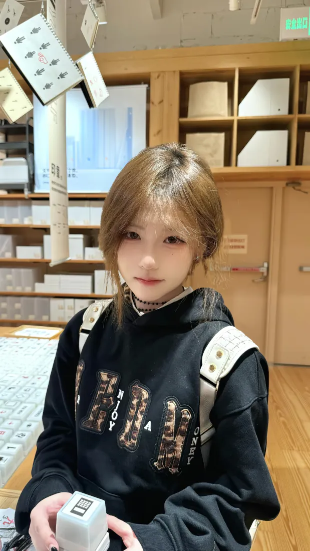 浅棕短发少女在木质手作店内自拍，身穿黑色连帽卫衣，手持白色印章。