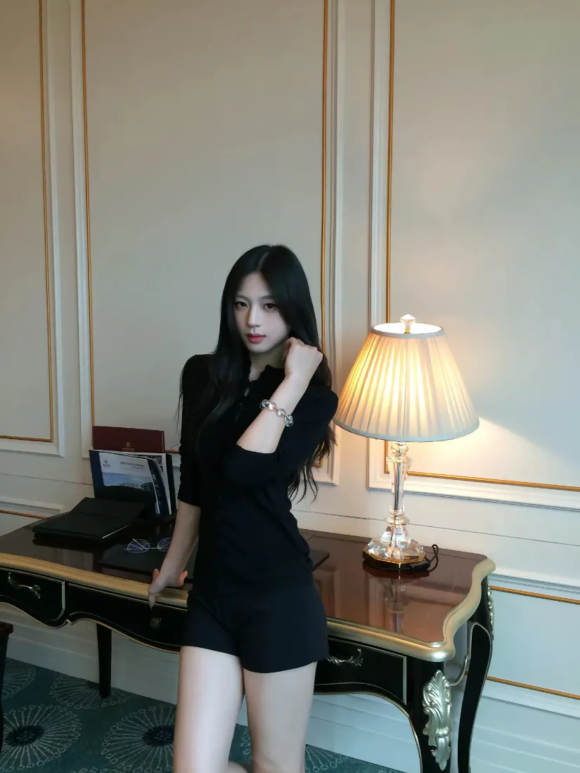 黑衣长发女子站在酒店书桌旁，身旁台灯点亮，背景是带金色线条的护墙板墙面。