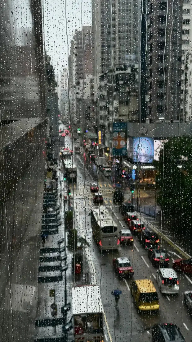透过布满雨滴的玻璃俯瞰雨天繁忙都市街道，车流穿梭，两侧高楼林立。