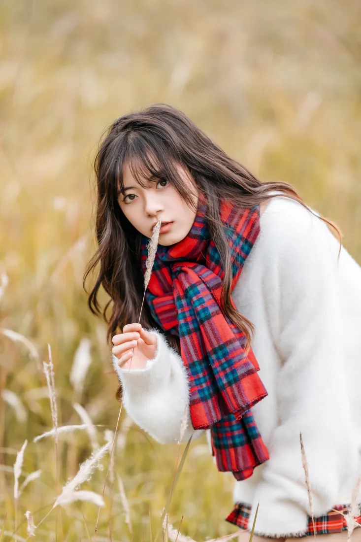 秋日户外芦苇丛中，穿白色毛衣戴格纹围巾的长发少女手持草穗望向镜头。