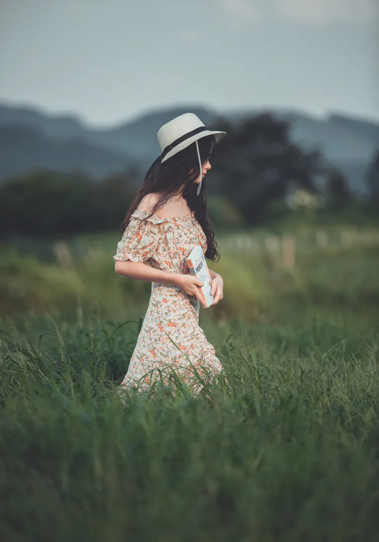 戴草帽的长发少女身着碎花露肩裙，手持书本站在郊野青草地中，背景远山朦胧。