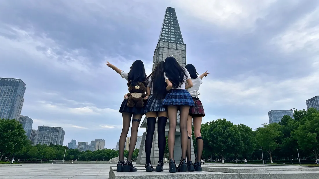 四名穿JK制服的长发少女背对镜头站在城市广场台阶上，身后是摩天大楼与绿树蓝天。
