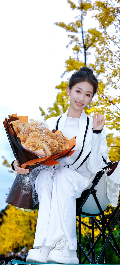 秋日户外，白衣丸子头女子坐折叠椅上，手持花束，身后是金黄银杏。