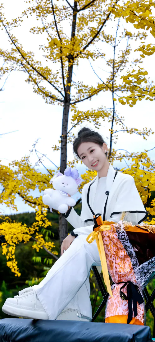 秋日户外，白衣丸子头女生坐于银杏树下，手持白色毛绒玩偶，身旁摆放花束。