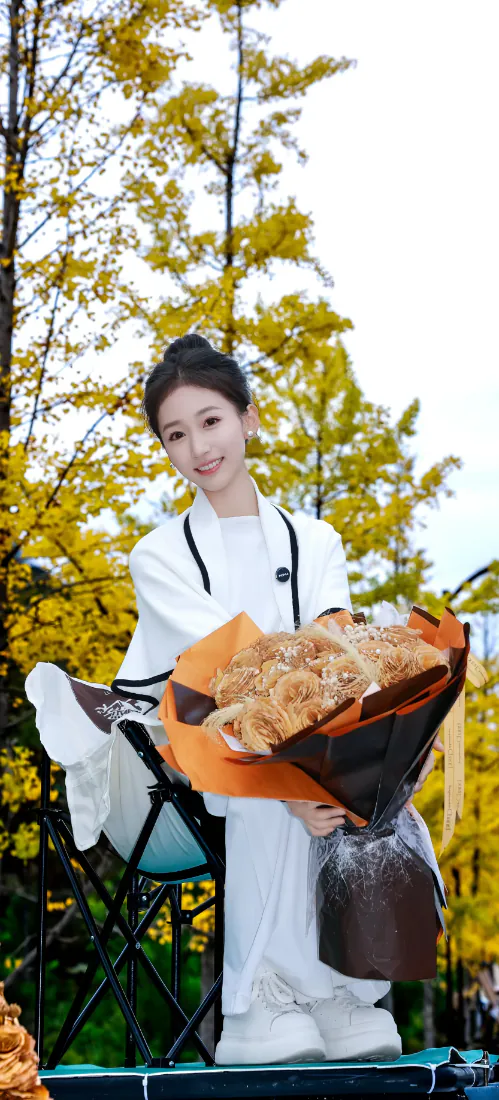 秋日银杏树下，白衣古风女子手捧花束，面带微笑站在户外。