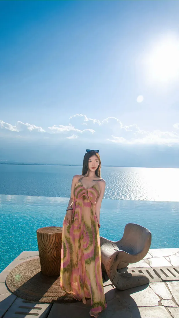 长发美女身着粉黄印花吊带长裙，站在临海无边泳池边的石质平台上，背景是蓝天碧海。