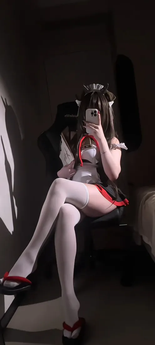 暗调室内，穿女仆装配白丝的女生坐椅上举手机对镜自拍，面部被手机遮挡。
