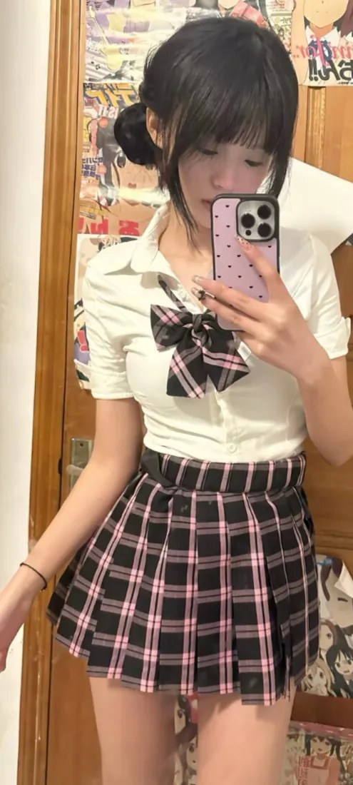 扎丸子头的少女穿JK制服，手持波点手机壳对镜自拍，背景木门贴满动漫海报。