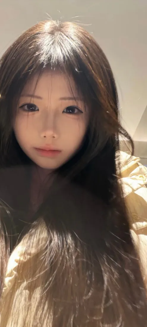 暖光下长发年轻女生近景自拍，背景为浅色室内墙面