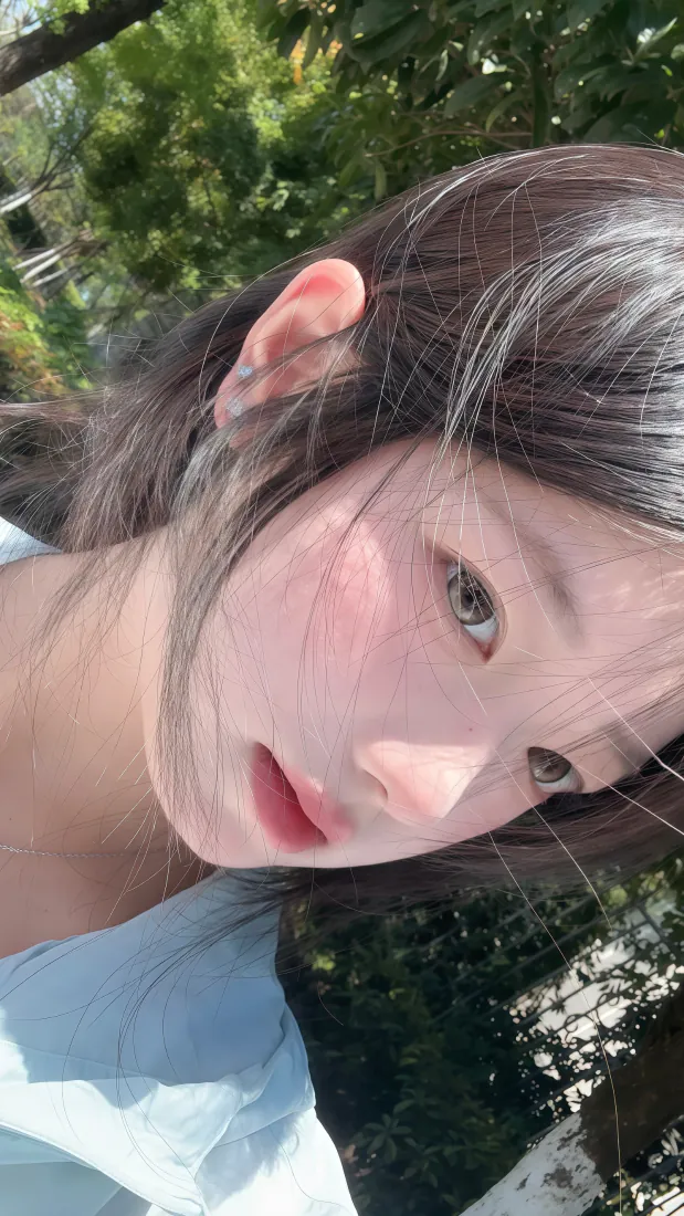 户外绿植旁的长发少女自拍，阳光洒落，身着浅蓝上衣，氛围清新治愈。