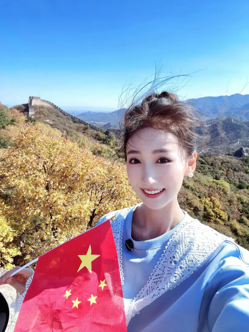 秋日晴朗白天，女子在长城上手持中国国旗自拍，背景是层叠山林与远山。