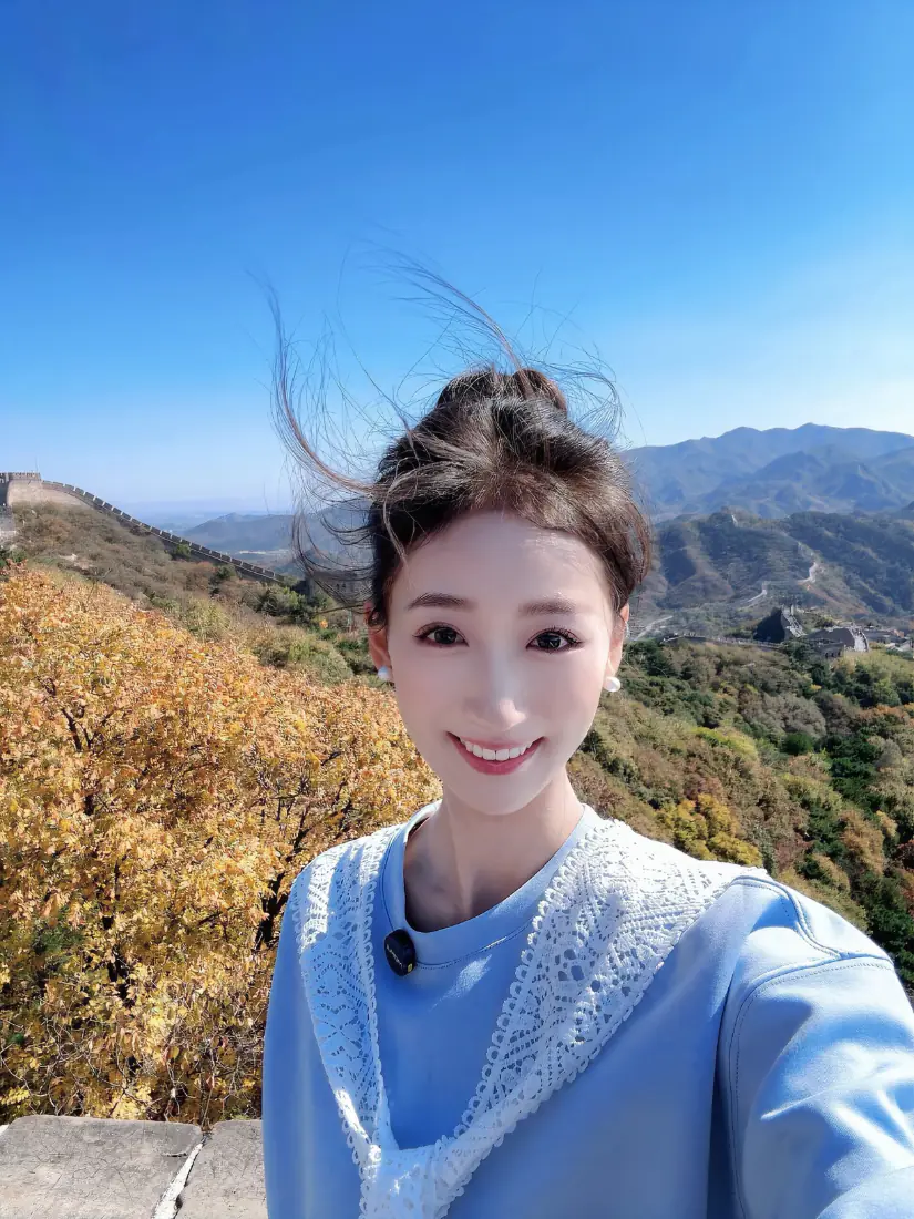 秋日长城上，盘发蓝衣美女微笑自拍，背景是蓝天秋林与远山长城。
