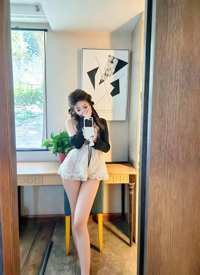 卷发少女在室内镜前自拍，穿白色蕾丝短裙搭配黑色长袖上衣，身后有书桌绿植与装饰画。
