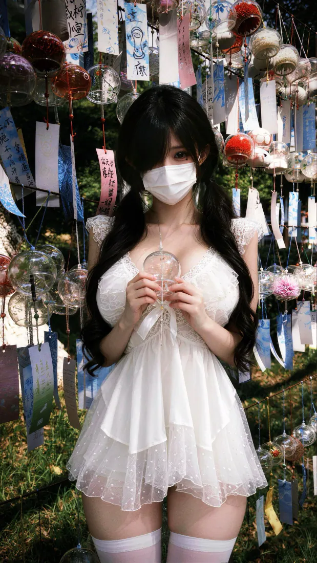 戴白色口罩的双马尾少女身着白纱裙站在挂满风铃的户外绿植前，双手持玻璃风铃。