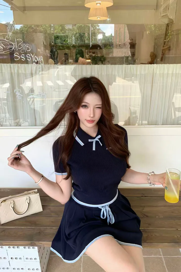 长卷发女生穿藏蓝连衣裙坐咖啡馆木桌旁，手持果汁眨眼微笑