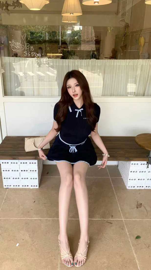 长发年轻女生身着藏青蝴蝶结短裙，坐在咖啡馆外木质长凳上，脚穿细带高跟凉鞋。