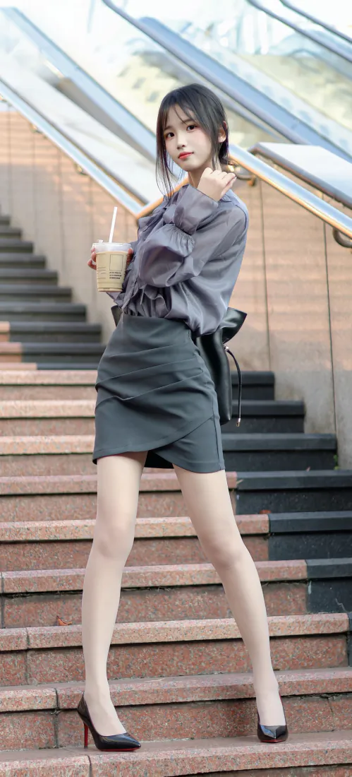 穿灰衬衫包臀裙的职场美女站在户外楼梯上，手持奶茶，脚踩红底高跟鞋。