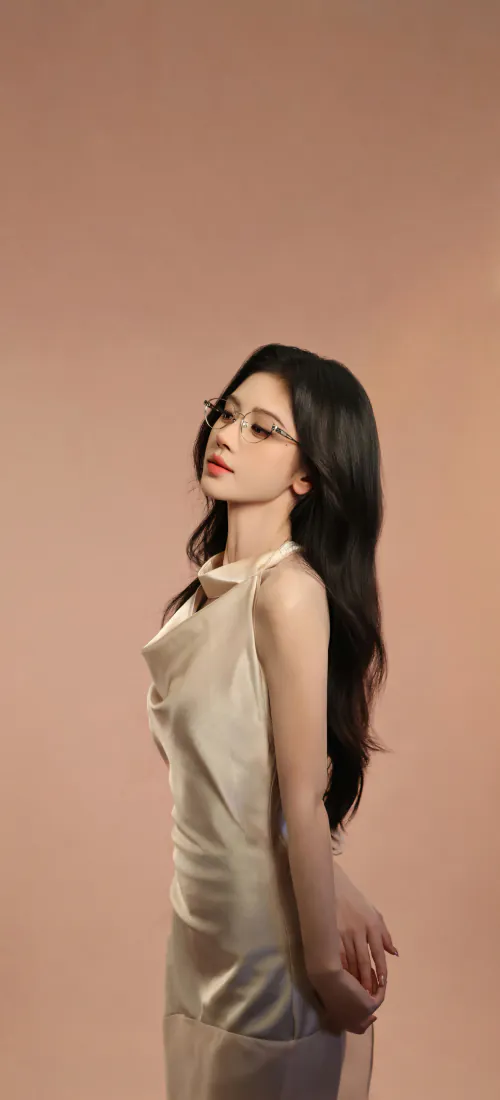 鞠婧祎戴细框眼镜，穿香槟色挂脖缎面裙，侧身立于暖棕纯色背景前。